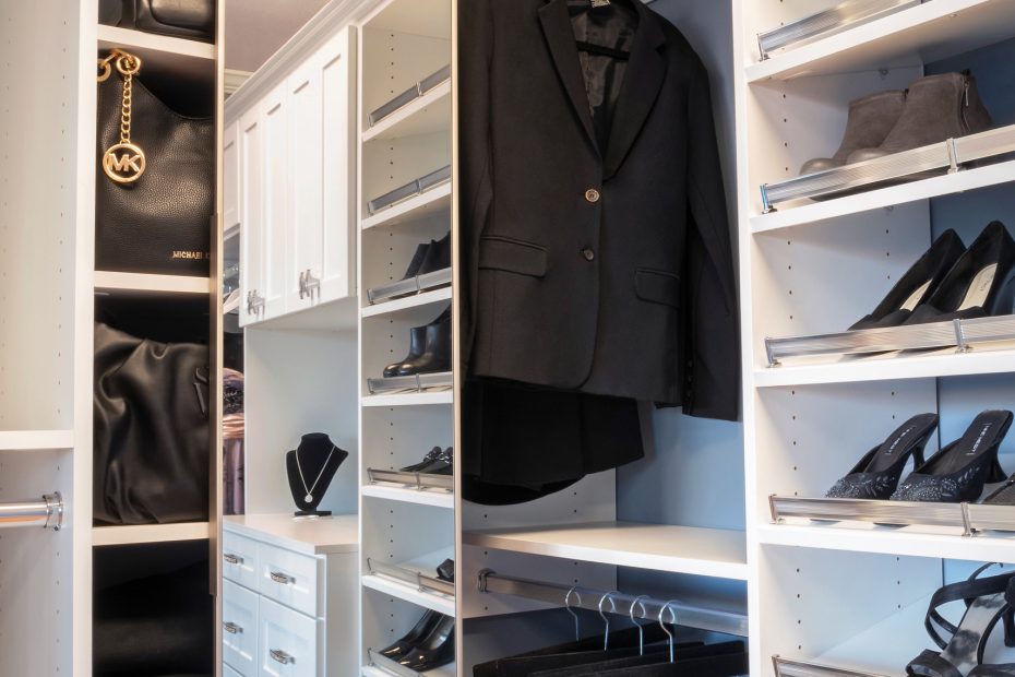 A neatly organized closet with a black suit hanging, shelves of shoes including heels, handbags, and drawers. The space is modern and elegant.