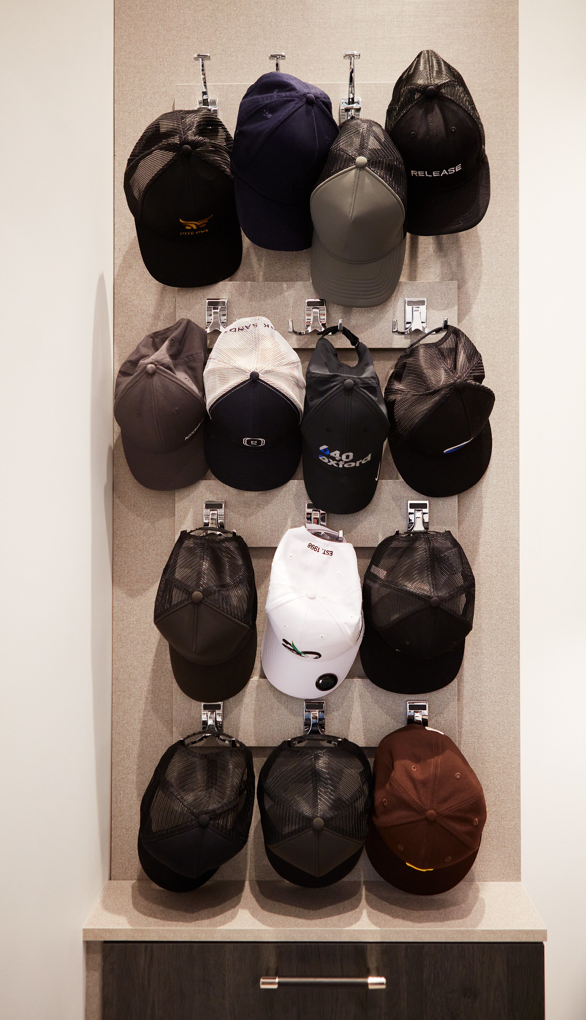 An impressive floor-to-ceiling custom hat wall display featuring twelve neatly organized baseball caps on individual hooks over a built-in lower storage cabinet.